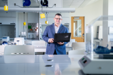 BIOCOMP alumnus Miguel Martinez stands in a laboratory holding an open laptop, surrounded by laboratory equipment and workstations.