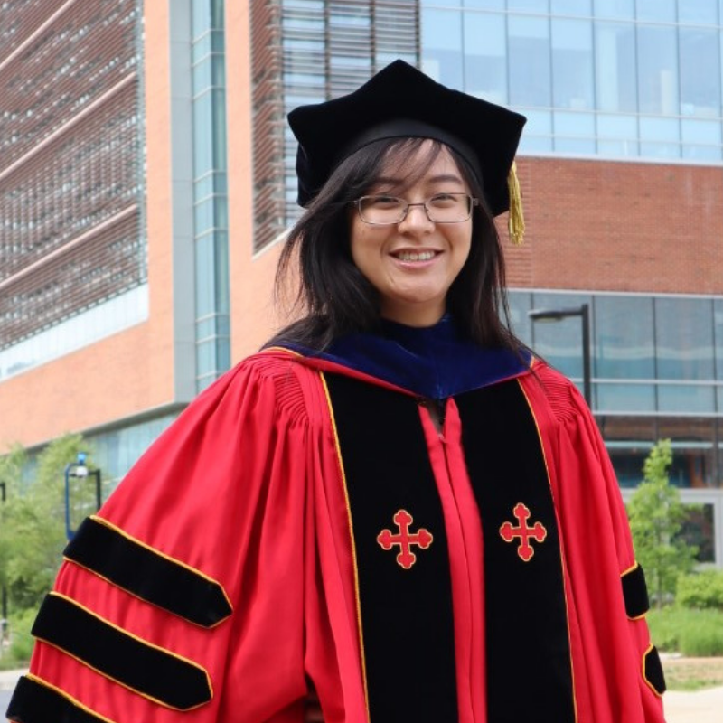 Photograph of Justine Yu in a red and black graduation cap and gown, smiling outside of Clark Hall